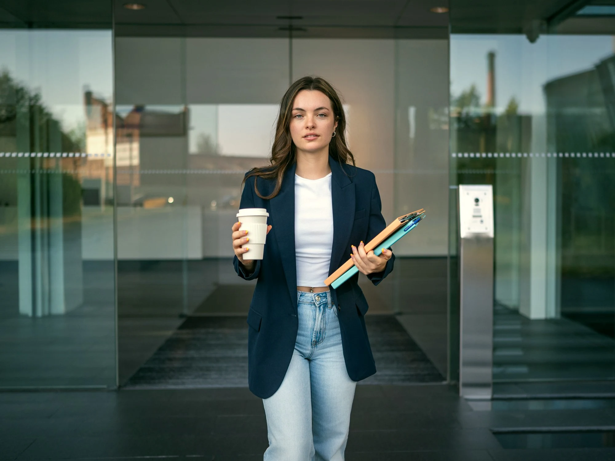 Unternehmerin mit Kaffeebecher und Unterlagen verlässt ein modernes Bürogebäude aus Glas